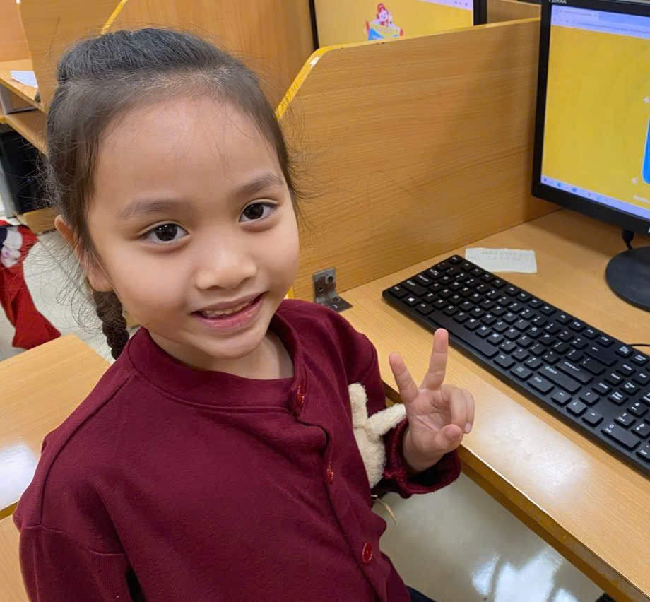 A child sitting at a desk with a computer and a hand gestureDescription automatically generated