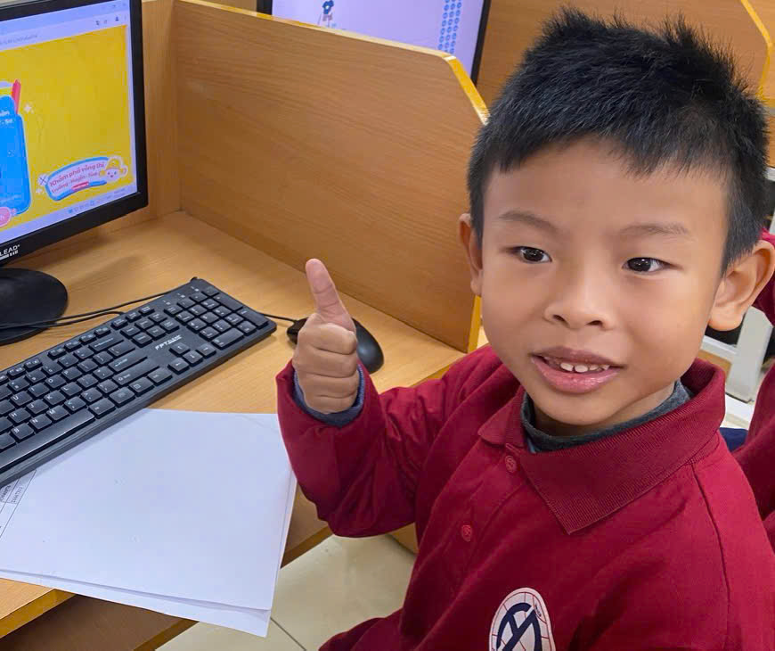 A child sitting at a desk with a computer and a thumb upDescription automatically generated