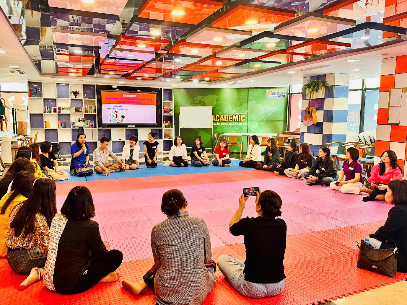 A group of people sitting on a mat in a room with a screen

AI-generated content may be incorrect.